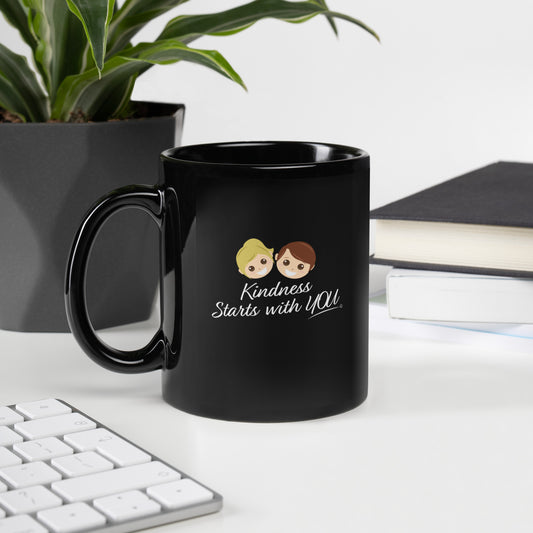 A 12-oz black mug featuring the uplifting quote 'Kindness Starts with You' with plant, keyboard and books in the background